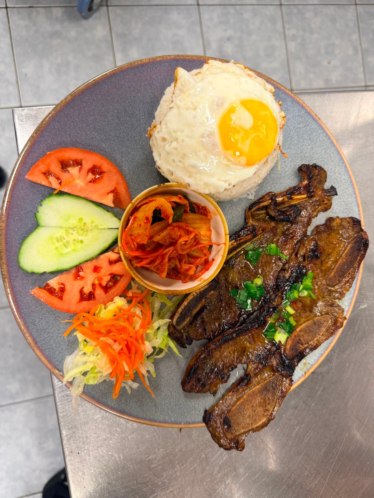 Grilled short ribs, rice topped with a fried egg, kimchi, and vegetables on a plate.